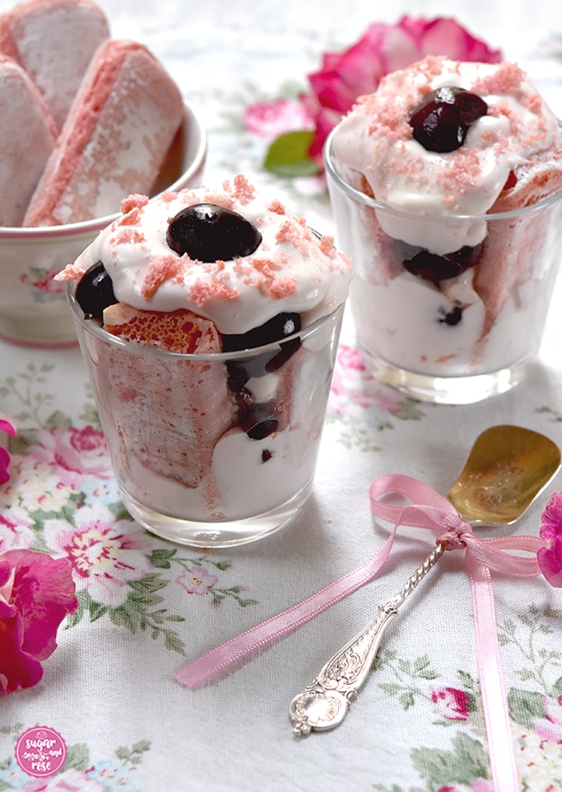 Großaufnahme von zwei Gläsern geschichtetes Kirschdessert auf einer mit rosa-grünen Rosenblüten dekorierten Leinenserviette. Im Vordergrund ein antiker Silberlöffel mit Gravuren
