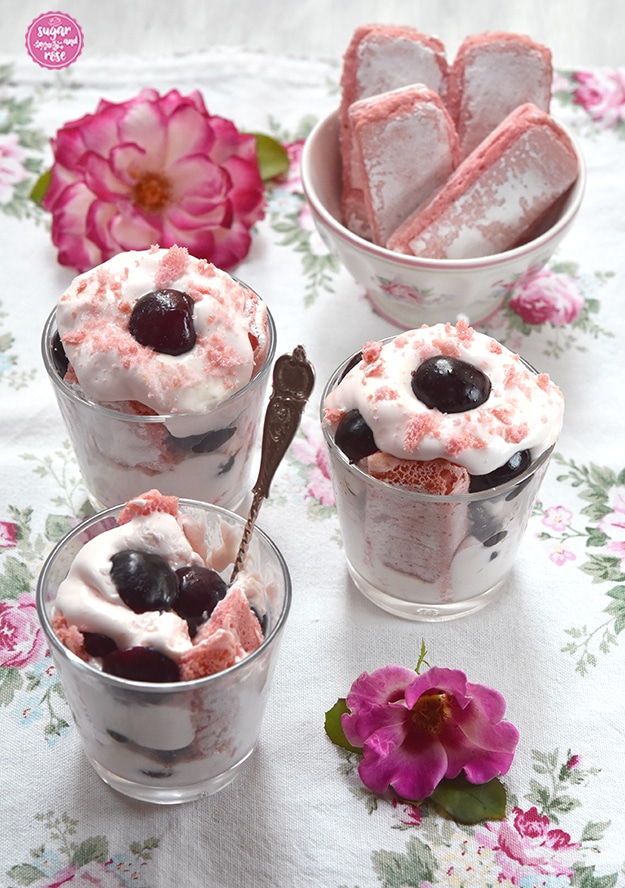 Drei Gläser Kirschdessert, im vorderen steckt der Silberlöffel im Glas. Es fehlt bereits ein Bissen. Im Hintergrund eine Schale mit vier Stück rosa Biskuits und rosa-weiße Rosenblüten