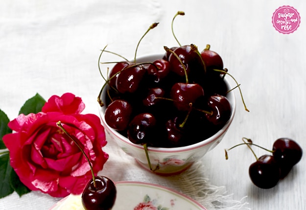 Eine kleine Porzeallanschale mit rosa Blüten gefüllt mit vollreifen dunkelroten Kirschen mit Stiel, daneben eine rot-weiß-marmorierte Rosenblüte