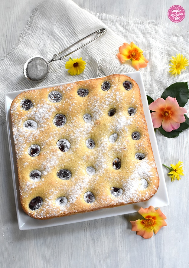 Obstkuchen mit Kirschen auf quadratischen weißen Teller, dahinter eine Zuckersieb und orange und gelbe offene Rosenblüten