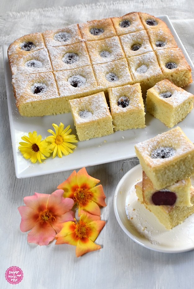 Ein quadratischer weißer Teller, darauf ein in kleine Quadrate geschnittener Kirschkuchen, davor ein kleiner weißer Teller, auf dem drei Stücke Kirschkuchen gestapelt sind, davor einige orange Rosenblüten.