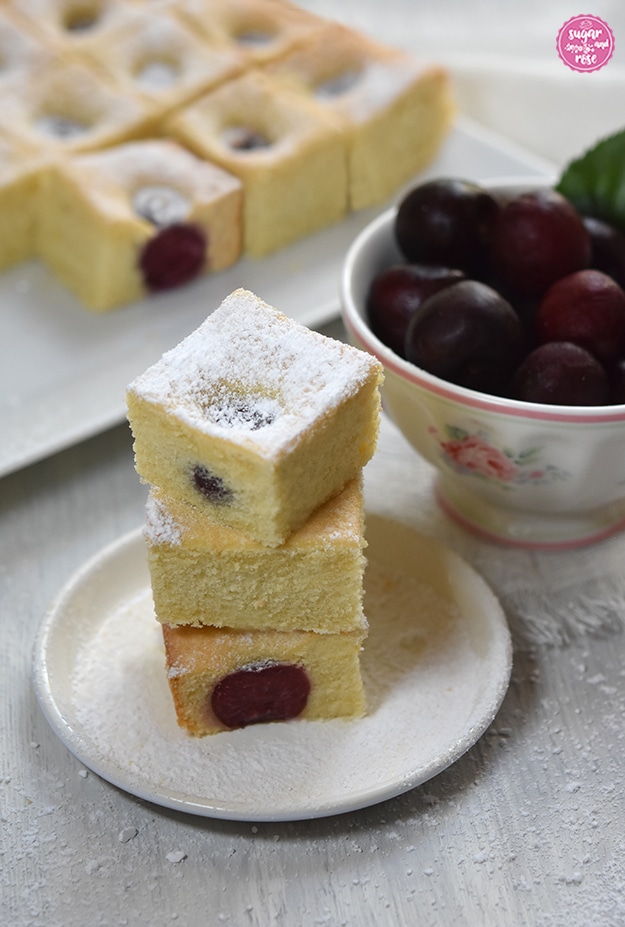Ein kleiner weißer Teller, darauf geschichtet drei Kirschkuchenstücke, dahinter eine kleine Porzeallanschale mit Kirschen und im Hintergrund der angeschnittene Obstkuchen mit Kirschen.