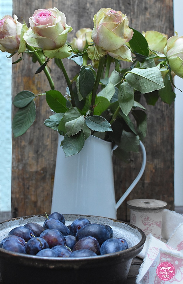 Eine braune Keramikschale mit kräftig blauen Zwetschken gefüllt, dahinter ein weißer Metallkrug mit blassrosa-grünen Rosen, daneben eine Holzspule mit breitem Leinenband