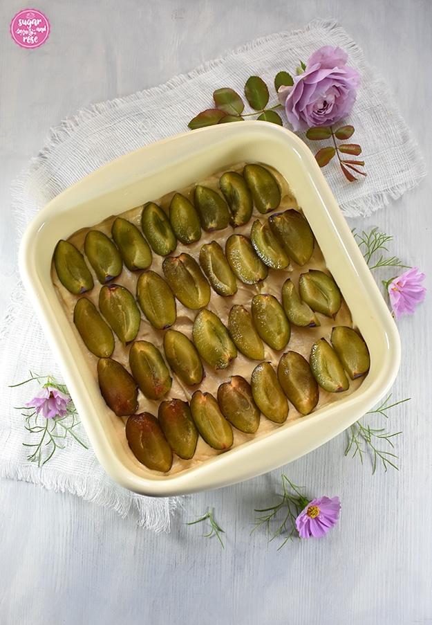 Quadratische Emailwanne mit noch ungebackenem Rührteig, darauf frische geviertelte Zwetschken mit Anschnitt nach oben. Lila Rose und Cosmeablüten als Deko rundum.