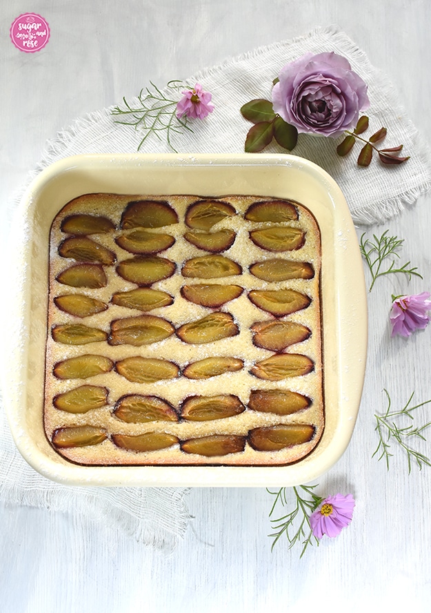 Obstkuchen mit Zwetschken in einer quadratischen Emailpfanne, dahinter eine lilafarbene Rosenblüte und einige rosa-lila farbene Cosmeablüten