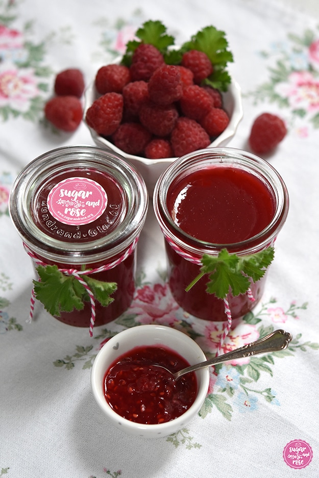 Zwei Gläser mit abgefülltem Himbeergelee, dekoriert mit rot-weißem Spagat und je einem grünen Pelargonienblatt. Ein Glas mit Deckel verschlossen, das andere offen. Davor eine kleine Schale mit Himbeermarmelade mit Kernen und einem Silberlöffelchen. Im Hintergrund eine Schale mit frischen Himbeeren, einige davon außerhalb am Geschirrtuch mit Rosenblüten liegend.