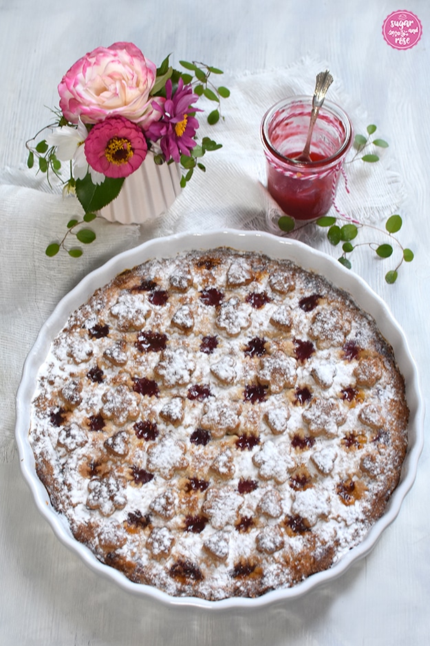 Linzer Torte in runder weißer Keramikform, mit Staubzucker bestäubt, dahinter eine kleine Vase mit einer weiß-rosa Rose, rosa Zinnien und grünen Blättern, daneben ein Glas Himbeergelee mit Silberlöffel