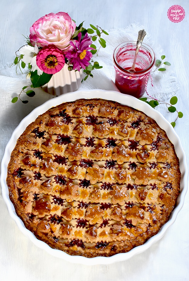 Frisch gebackene Linzer Torte in runder weißer Keramikform, ungezuckert, dahinter eine kleine Vase mit rosa Blüten und grünen Blättern, daneben ein Glas Himbeergelee mit Silberlöffel