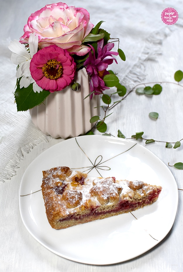 Ein Stück der Linzer Torte auf einem weißen Keramikdessertteller mit zarter goldener Masche als Dekor, im Hintergrund eine dezent rosa Vase mit Rose, Zinnie und weißen und lila Cosmeablüten, davor kleine grüne Blätter liegend