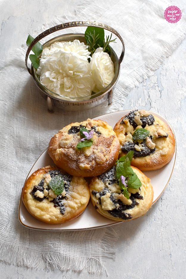 Vier Kolatschen auf einem ovalen Porzellanteller, dahinter eine kleine silberne Art-Deco-Schale mit Henkel, gefüllt mit drei weißen Rosenblüten.