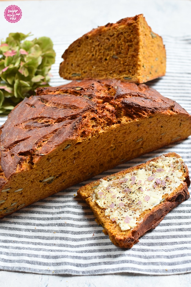 Halber Laib Kürbisbrot, dahinter ein Viertellaib und davor eine Scheibe Brot mit Butter und grobem rosafarbenen Salz