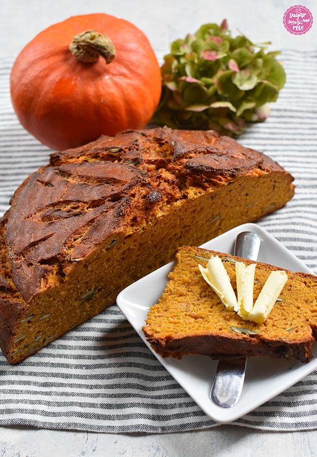 Halbierter Kürbisbrot-Laib mit Hokkaidokürbis und Hortensienblüte im Hintergrund; vorne ein kleiner quadratischer weißer Teller mit einer Scheibe Brot, drei Röllchen Butter und ein kleines Buttermesser