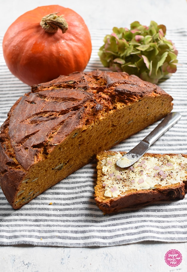 Ein halber Laib Kürbisbrot, dahinter ein Viertellaib und davor eine Scheibe Brot mit Butter und grobem rosafarbenen Salz und einem Buttermesser