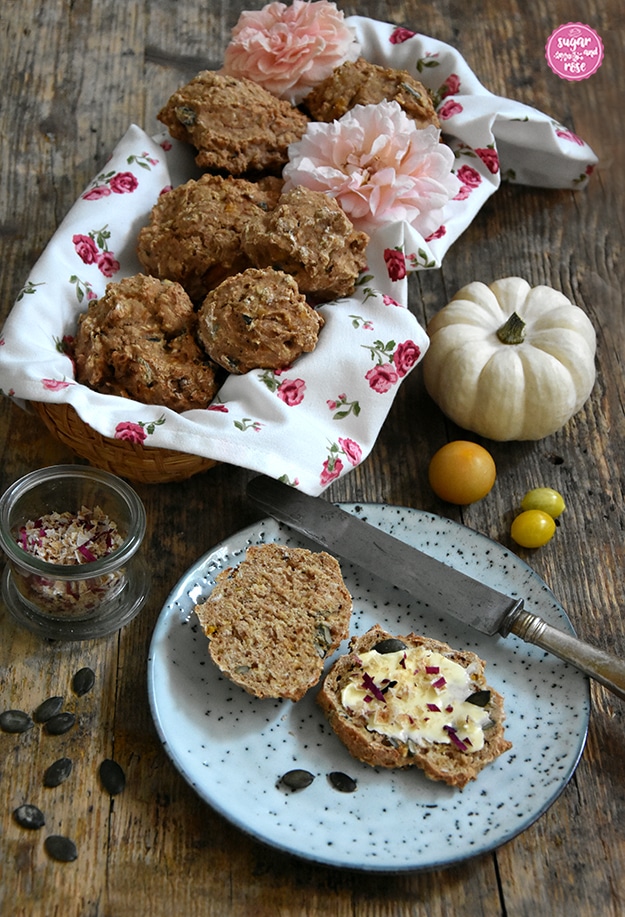 Mehrere Vollkornweckerl in einem Brotkörbchen mit Geschirrtuch mit kleine Rosenblüten, davor ein grauer Keramikteller mit einem aufgeschnittenen Brötchen, bestrichen mit Butter, bestreut mit Kürbiskernen, grobem Rosensalz, daneben ein altes Messer und kleine gelbe Tomaten