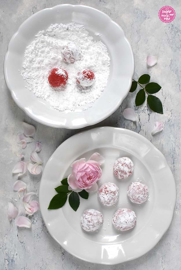 Zwei weiße Keramikteller, im hinteren Staubzucker mit drei rosa Teigbällchen, davor fünf bereits in Zucker gewälzte Cookieteigkugeln.