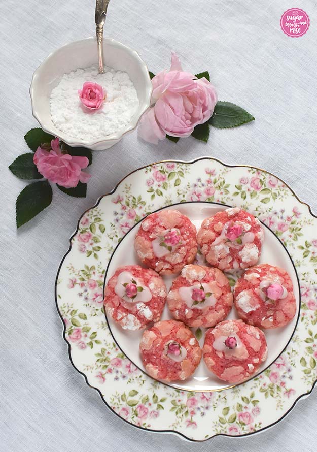 Ein weißer Porzellandesserteller mit Goldrand und dichtem Rosendekor am Tellerrand - gefüllt mit sieben im Kreis aufgelegten Rosen-Cookies. Als Deko dahinter wieder eine Zuckerschale und rosa Rosenblüten.