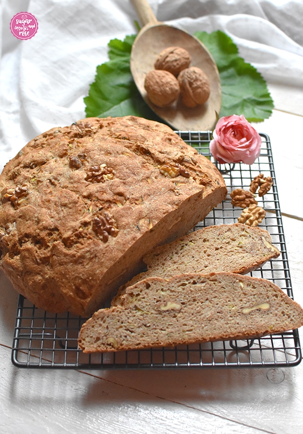 Ein angeschnittener Laib Walnussbrot und zwei Brotscheiben sowie Walnusskerne auf einem grauen Gitterrost, dahinter ein kleine hölzerne Mehlschaufel mit ganzen Walnüssen