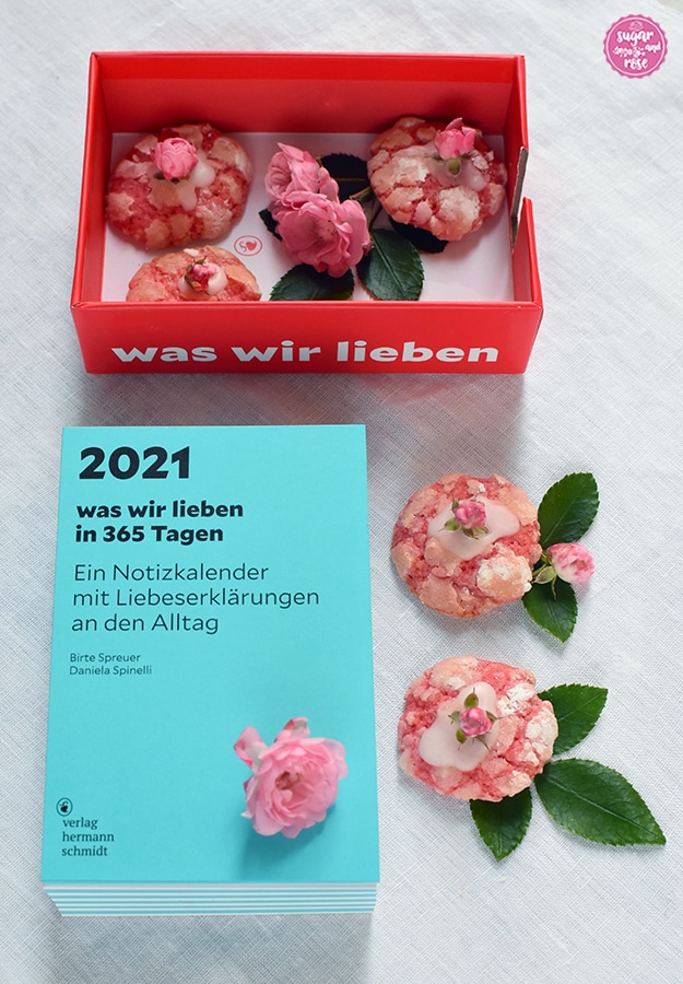 Im Vordergrund ein hellblauer Stapel von Kalenderblättern (Kalenderblock), darauf ein kleines Röschen, daneben zwei Rosen-Cookies mit kleinen Rosenblüten dekoriert; im Hintergrund die rote Kartonschachtel, mit der der Notizkalender ausgeliefert wird. In der Schachtel liegen Rosenkekse und Rosenblüten.