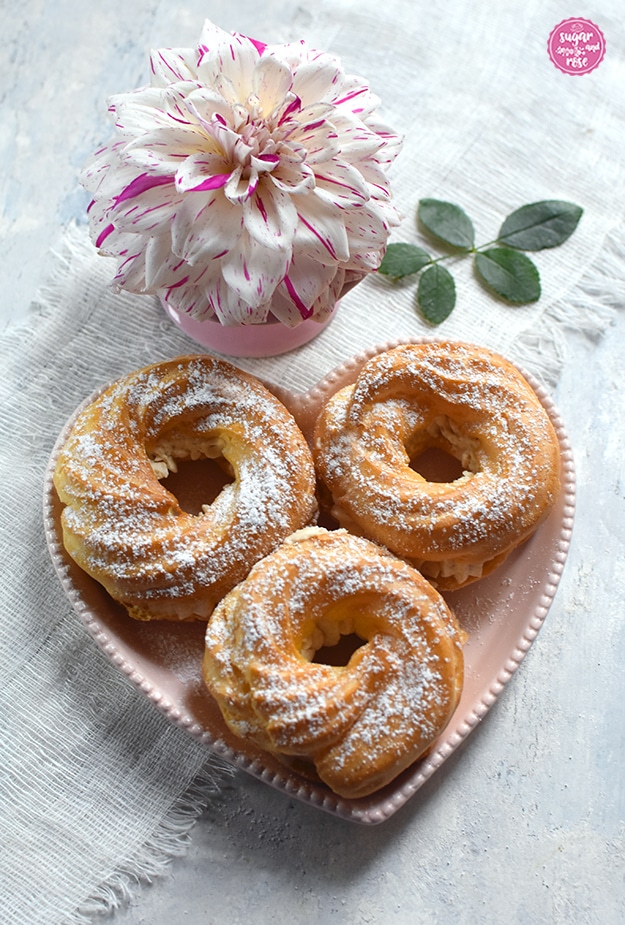 Drei gefüllte Brandteigringe in einem herzförmigen rosa Teller, dahinter eine pink-weiß gesprenkelte Dahlienblüte