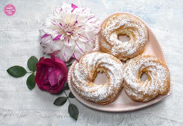 Im Querformat: ein dezent rosa Herzteller mit gefüllten und mit Staubzucker bestreuten Brandteigringen, links daneben eine weiß-pinke Dahlienblüte, eine dunkelpinke Rosenblüte und grüne Rosenblätter