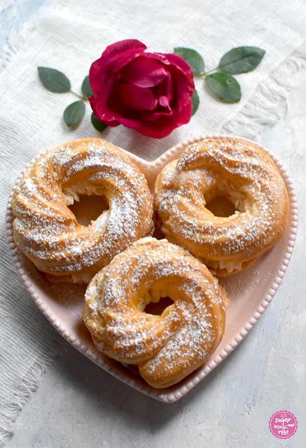 Drei gefüllte Brandteigringe in einem herzförmigen rosa Teller, dahinter eine dunkelpinke Malerrosenblüte