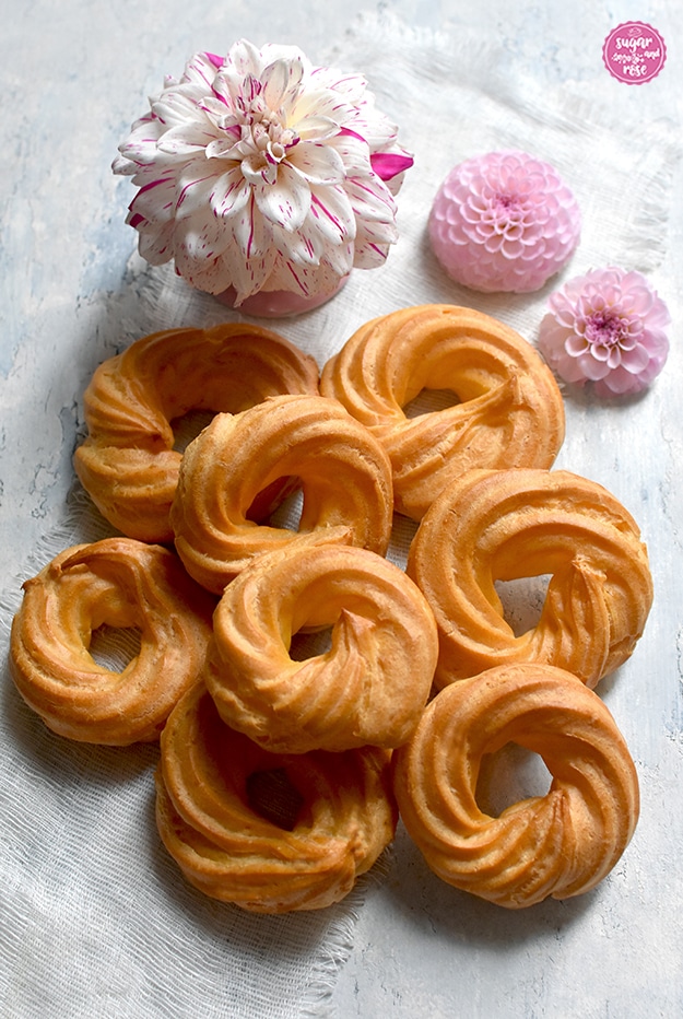 Acht Stück ungefüllte Brandteigringe übereinander liegend, daneben zartrosa Dahlienblüten und eine weiß-pinke Dahlienblüte auf einer winzigen rosa Porzellanetagere