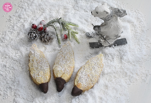 Drei Tannenzapfen-Kekse auf Kunstschnee, dahinter ein  Deko-Eisbär auf Schiern, ein grüner Zweig, weiß beschneite Bockerl und rote Kugerl