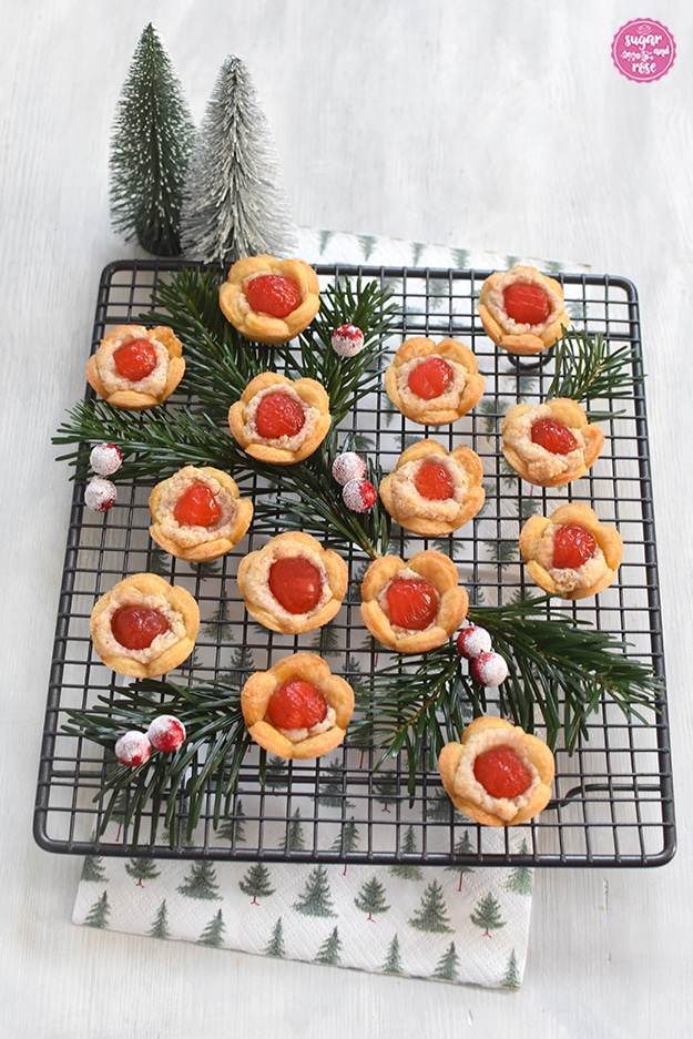 Christrosen-Kekse auf einem dunklen quadratischen Gitterrost mit Tannenzweigen und roten Früchten dekoriert, darunter eine Serviette mit Tannenbaummotiven und im Hintergrund zwei grüne Deko-Tannenbäumchen