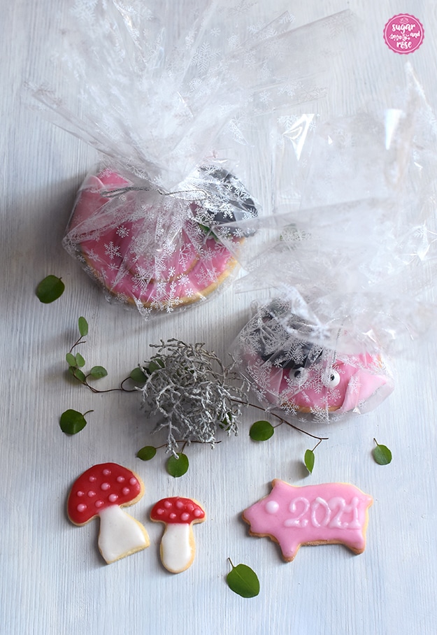 Zwei Glücksschweinchen-Kekse verpackt in durchsichtiges Cellophan mit weißen Kristallen drauf, davor zwei Glückspilz-Kekse und ein rosa glasiertes Keks-Schweinchen mit der Aufschrift in weißer Zuckerglasur "2021"