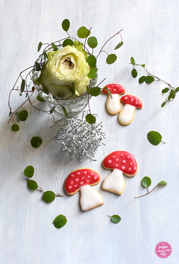 Zwei große und zwei kleine Glückspilz-Kekse mit roter und weißer Zuckerglasur, dahinter ein kleines Bleikristallväschen mit einer grün-weißen Rosenblüte und grünen Blättern