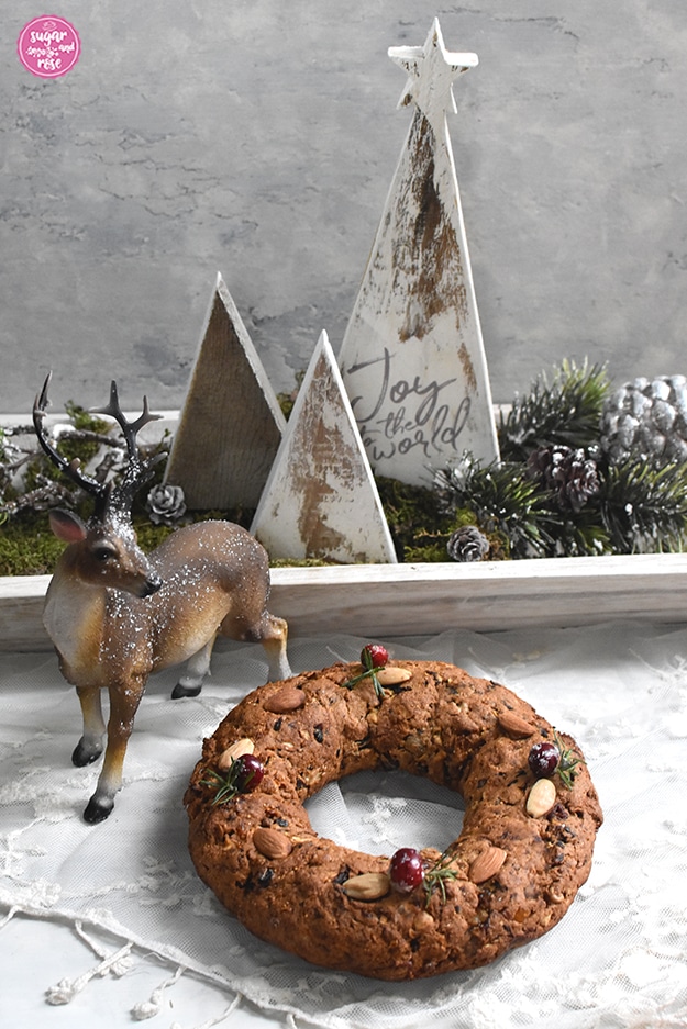 Kletzen-Früchtebrot in Ringform, dekoriert mit Cranberrys, Rosmarin und Mandeln vor einer weißen Holzschale mit Holzbäumen, Reisig, Zapfen und einer Hirschfigur