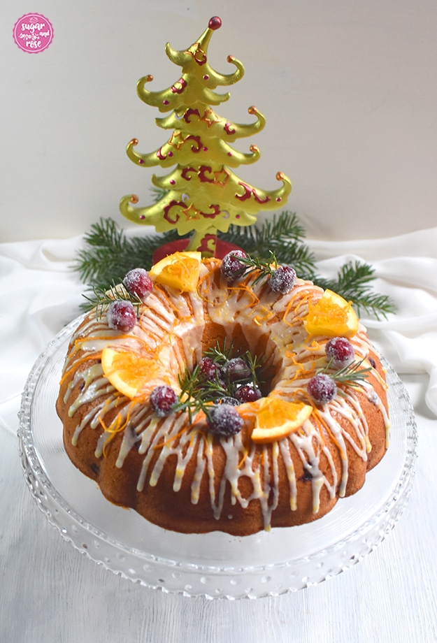 Orangen-Cranberry-Gugel auf einem gläsernen Kuchenteller mit Standfuß, dahinter ein grün-roter Weihnachtsbaum aus Keramik und Tannenzweige