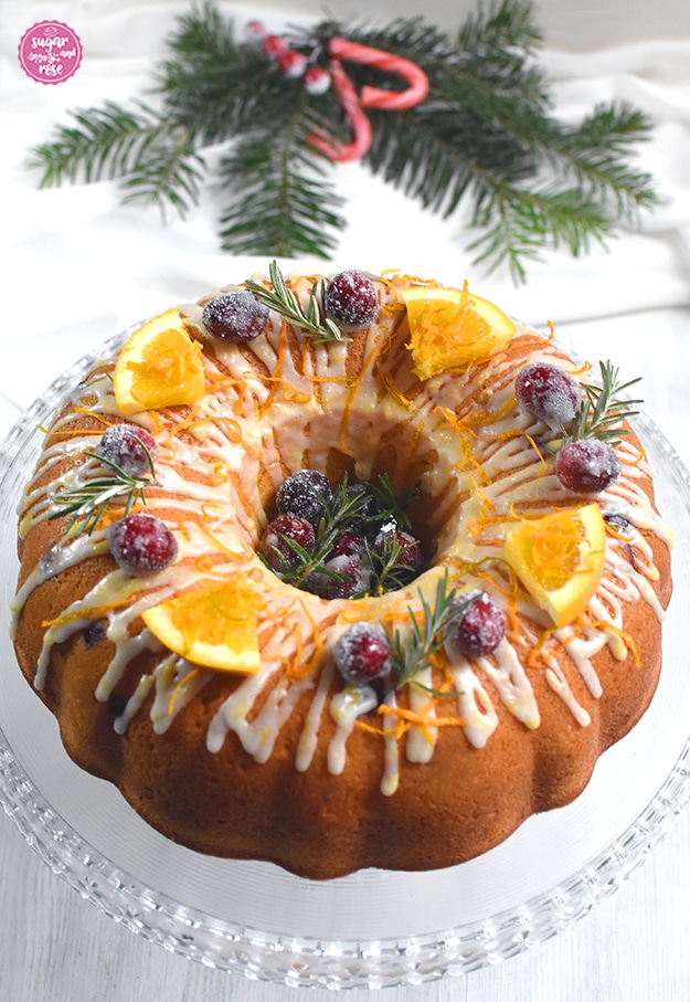Orangen-Cranberry-Kranz auf gläserner Etagere mit gezuckerten Cranberrys in der Mitte und als Deko, dahinter Tannenzweige und rote Zuckerstangen
