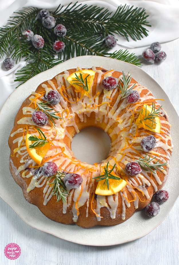 Orangen-Cranberry-Kranz mit Orangen-Icing, dekoriert mit gezuckerten Cranberrys und Orangenspalten sowie grünen Rosmarinzweigen auf einem rohweißen unregelmäßigen Keramikteller