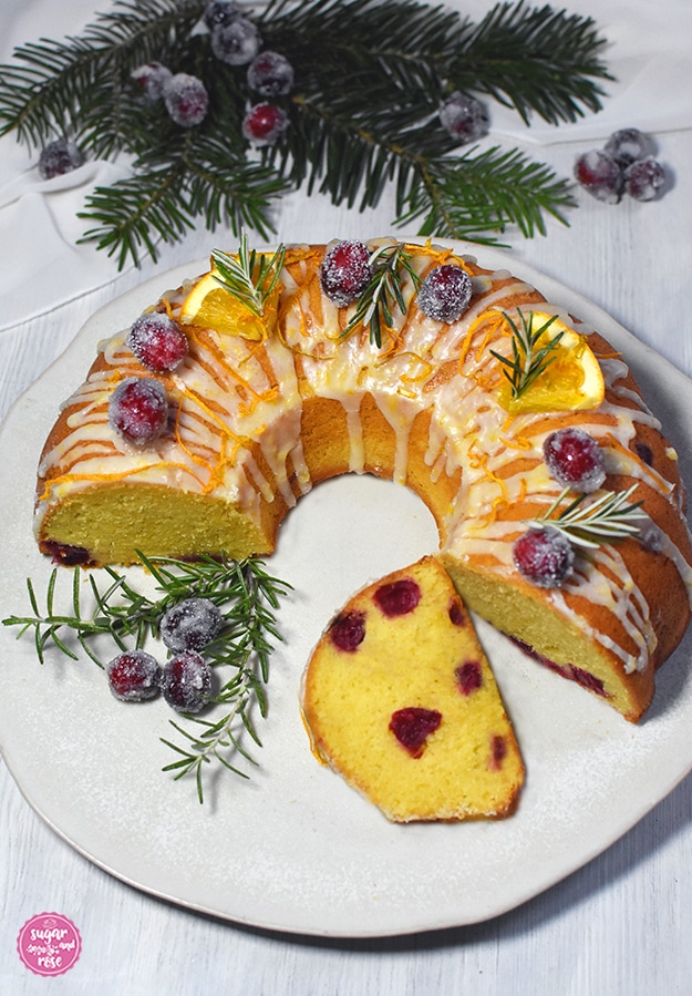 Angeschnittener, halber Orangen-Cranberry-Kranz, davor ein Stück vom Kuchen, saftig, knallgelb und mit zahlreichen roten Cranberry-Einschlüssen, dahinter Tannenzweige und gezuckerte Cranberrys