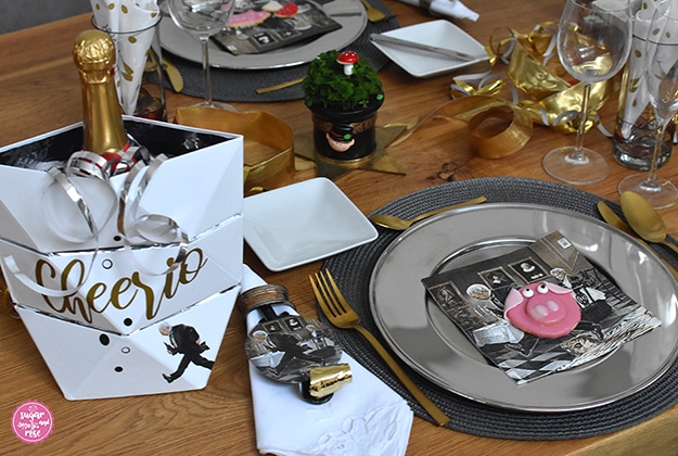 Ein Gedeck mit Silber-Platzteller und Goldbesteck, einer Papierserviette mit Motiv "Dinner for one" und Glückschweinchen-Kekse am Teller