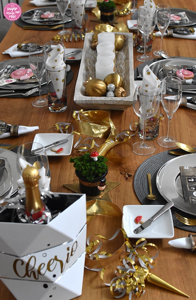 Tischdeko mit Glücksklee und Sektflasche in einem weißen Kühler mit der Aufschrift "Cheerio".