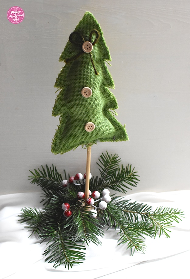 Selbstgenähter Weihnachtsbaum aus mittelgrünem Grobleinen auf einem Holzstab, dekoriert mit drei Holzknöpfen und einer grünen Masche, stehend auf Tannenzweigen mit roten kleinen Beerenfrüchten