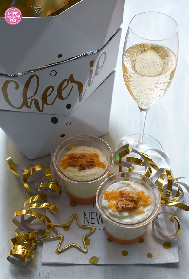 Zwei Dessertgläschen mit Karamell-Mousse, Goldgirlanden, ein Sektkühler aus weißem Karton mit der Aufschrift "Cheerio", daneben ein Glas Sekt