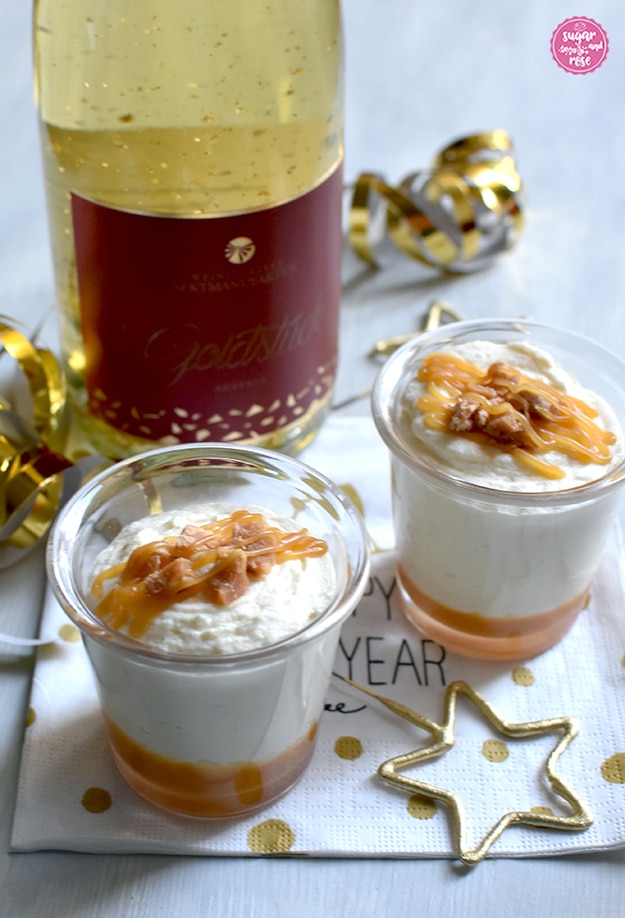 Zwei Gläschen mit Karamell-Mousse auf goldgepunkteter Papierserviette, im Vordergrund eine goldene sternförmige Spritzkerze, im Hintergrund eine Flasche Sekt mit Goldflitter (Goldsekt)