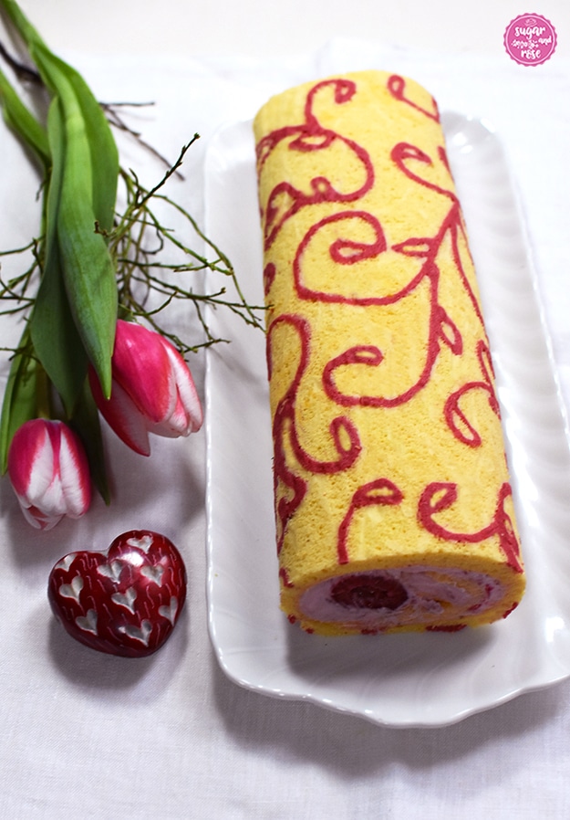 Motiv-Biskuitroulade auf weißem Porzellankuchenteller, daneben zwei rosa-weiße Tulpen und ein rotes Steinherz mit eingravierten kleinen weißen Herzen