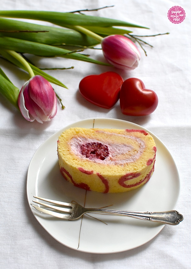 Ein Stück der mit rosa Topfencreme und Himbeere gefüllten Motiv-Bikuitroulade auf einem kleinen Porzellanteller, darauf auch eine kleine silberne Dessergabel. Im Hintergrund ein Stauß rosa Tulpen und zwei rote Steinherzen