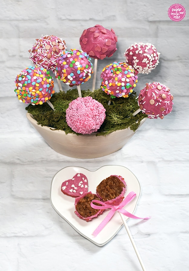 Acht rosa Cake-Pops in moosbedeckter Keramikschale, davor ein kleiner herzförmiger Keramikteller in weiß mit zartem grauem Rand, darauf ein halbierter Cake-Pop mit rosa Schleife. Die braune Punschfüllung ist sichtbar