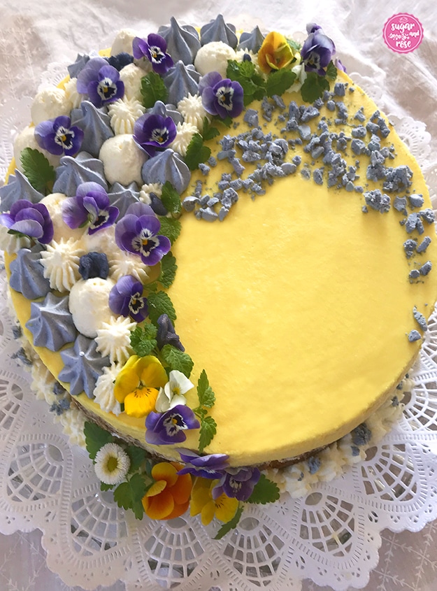 Satt gelbe Eierlikör-Torte von oben, zur Hälfte dekoriert mit leuchtend violetten Stiefmütterchen, blassvioletten Baisertuffs, weißen Cremetupfen und einigen zerbröselten Baiserstücken.