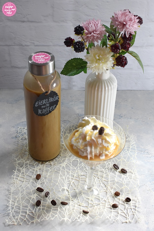 Eierlikör abgefüllt in Glasflasche, davor ein Cocktailglas mit Kaffee-Eierlikör und einer großen Sahnehaube, daneben eine weiße kleine Porzeallanvase in Flaschenform mit rosa und weißen Kunstblüten sowie Brombeeren