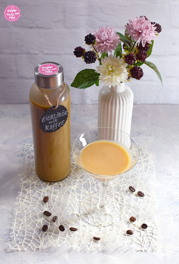 Kaffee-Eierlikör abgefüllt in Glasflasche mit beschriftetem, rundem schwarzen Etikett, am Deckel der Flasche ein rosa Kleber, davor ein Cocktailglas mit Kaffee-Eierlikör, daneben eine weiße kleine Porzeallanvase in Flaschenform mit rosa und weißen Kunstblüten sowie Brombeeren