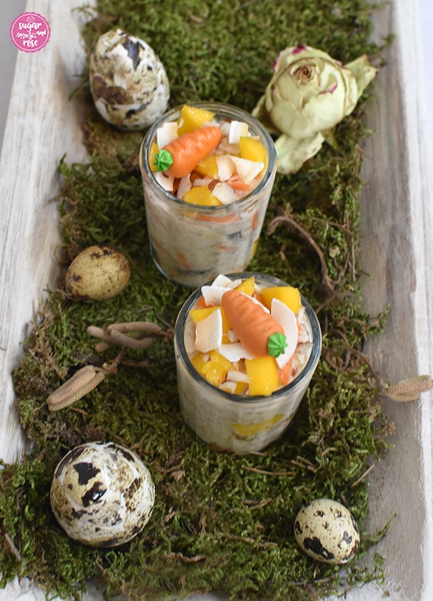 Zwei kleine Gläser mit Karotten-Overnight-Oats, dekoriert mit je einer Marzipan-Karotte. Die Gläser stehen auf einer hellen länglichen Holzschale mit Moos, dekoriert mit Wachteleiern