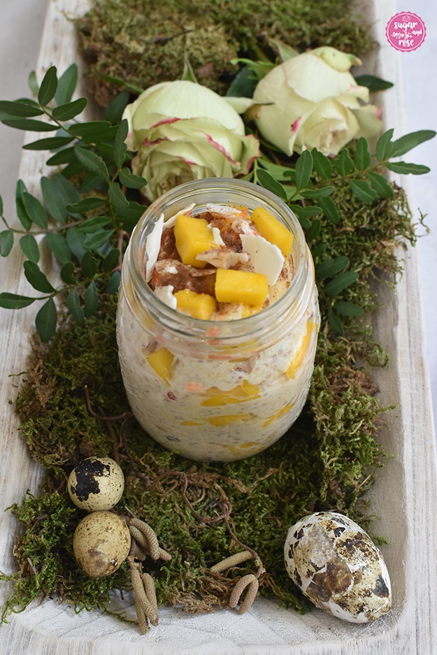 Karotten-Overnight-Oats im Schraubglas auf grünem Moos, dahinter zwei grün-weiße Rosen mit zartrosa Rand