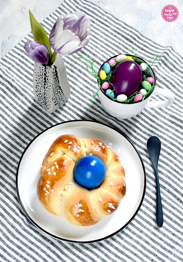 Im VordergRund ein weißer Teller mit dunkelblauem Rand, darauf ein Brioche-Osternest mit Hagelzucker und blauem Ei in der Mitte, daneben ein schwarzes Löffelchen, dahinter eine kleine graue Vase mit lila Tulpenblüten, daneben ein Häferl mit einem violetten Ei und rundum kleinen bunten Zuckereiern