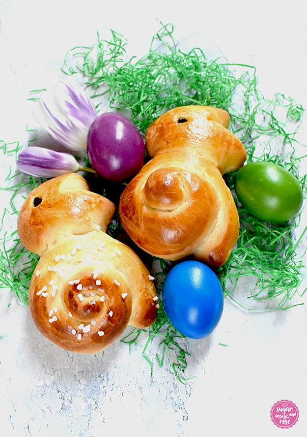 Zwei Brioche-Osterhasen auf grünem Papierstroh, je ein violettes, grünes und mittelblaues Ei und zwei violett-weiße Tulpenblüten